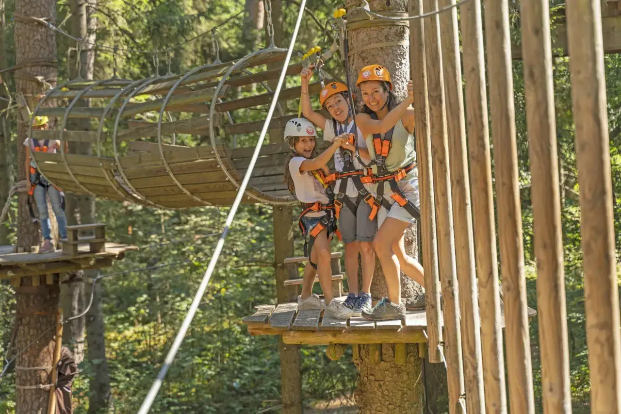 Familie genießt das Klettern im Hochseilgarten. Kinder und Mutter mit Helmen und Gurten auf einer Plattform, bereit für das nächste Abenteuer.