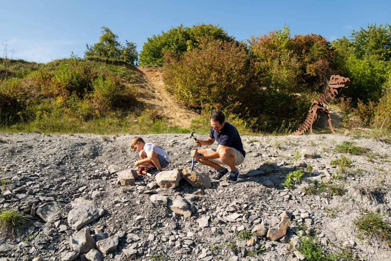 Ein Vater und sein Sohn graben nach Fossilien. Im Hintergrund steht ein Skelett-Dinosaurier aus Metall.
