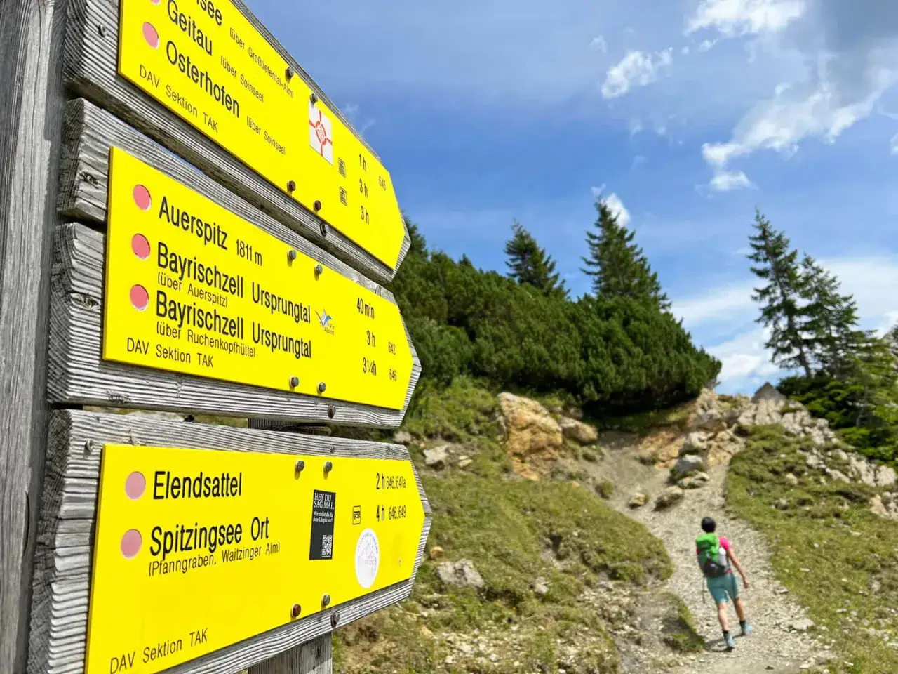 Wegweiser für bayrischzell wandern mit kindern: Auerspitz, Ursprungtal, Elendsattel. Ein Kind mit Rucksack steigt den Bergpfad hinauf.