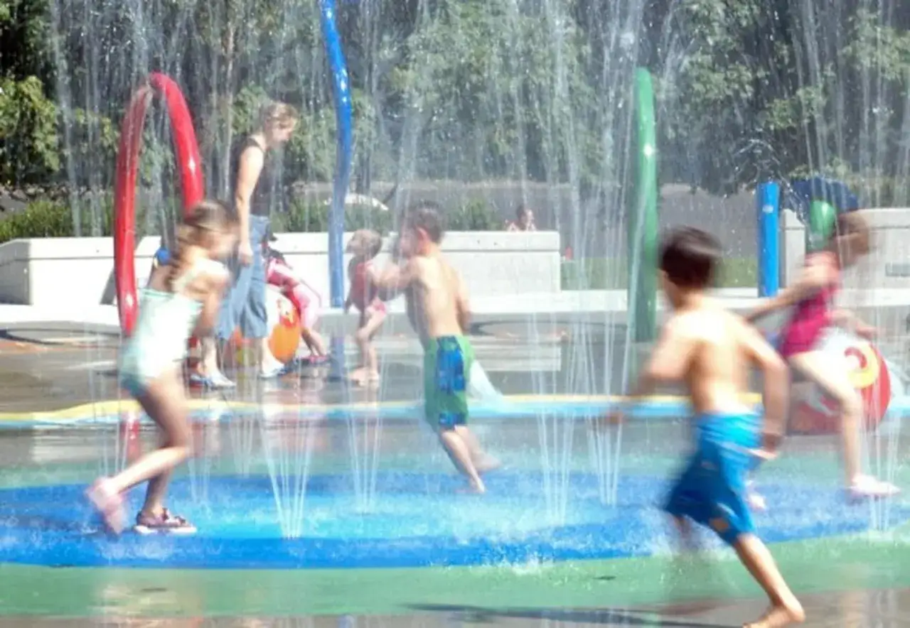Kinder planschen und lachen auf einem bunten Wasserspielplatz, w&auml;hrend Wasserfont&auml;nen sie erfrischen.