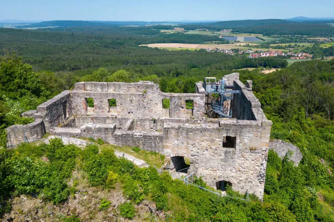 Die Ruine Bramberg thront auf einem bewaldeten Hügel. Steinmauern mit Fensteröffnungen und eine Wendeltreppe sind sichtbar. Im Hintergrund erstreckt sich eine grüne Landschaft mit Feldern und einem Dorf.