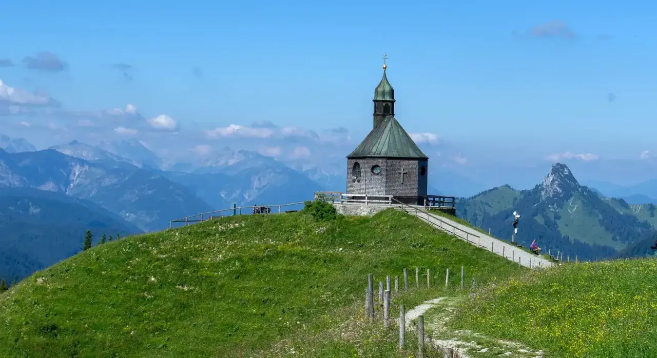 Altes Wallberghaus Wanderung: Kleine Kapelle auf einem grünen Hügel mit Bergpanorama. Ein Weg führt zur Kapelle, die Wanderer zur Rast einlädt.