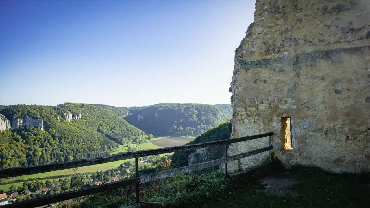 Blick von der Ruine Kallenberg &uuml;ber ein gr&uuml;nes Tal mit Felsen und W&auml;ldern. Ein schmaler Lichtschacht erhellt die alte Mauer.