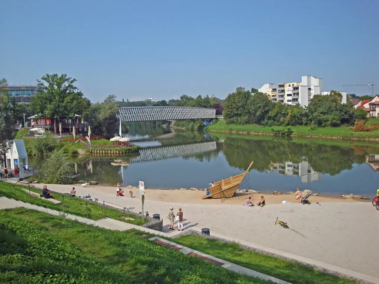 Sonniger Tag am Neckarstrand Remseck mit Menschen am Sandstrand, einem Spielschiff und einer Br&uuml;cke &uuml;ber dem Fluss.