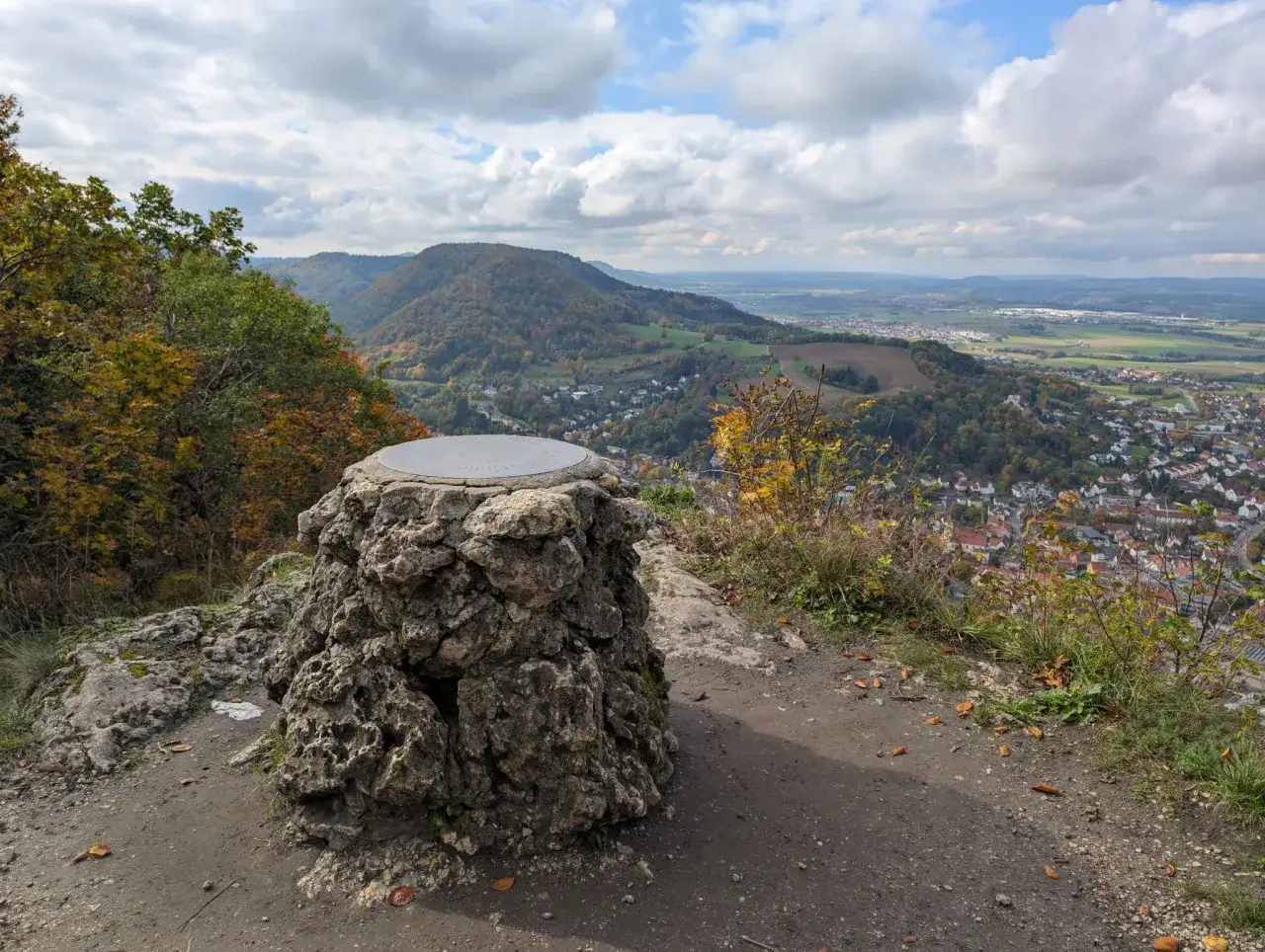 Blick vom Gipfelkreuz auf Heubach. Ein steinernes Monument mit einer Metallplatte steht im Vordergrund, w&auml;hrend sich im Tal die Stadt und bewaldete H&uuml;gel erstrecken. Perfekt f&uuml;r alle, die gerne heubach wandern.