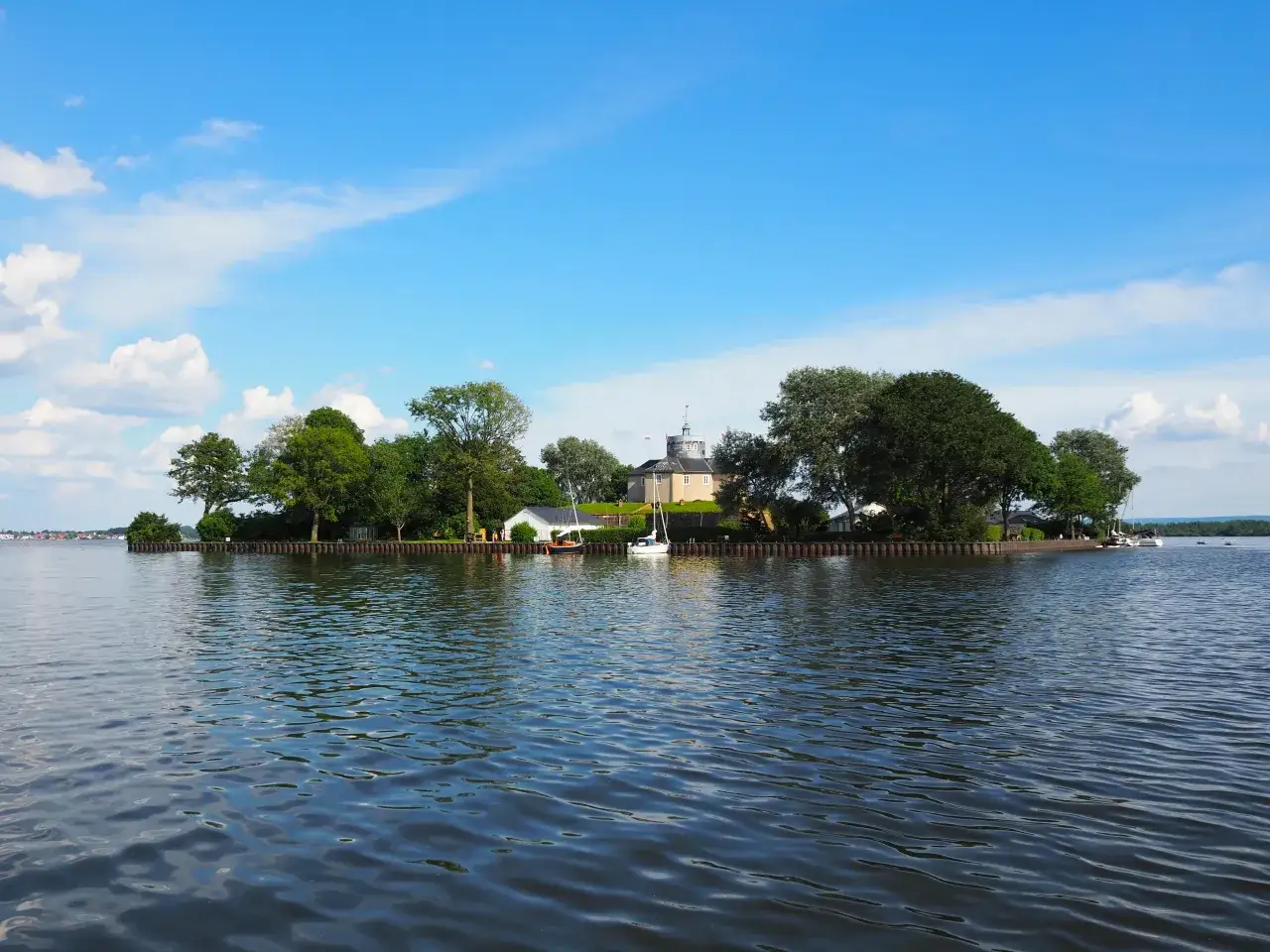 Insel mit Schloss und Bäumen am Steinhuder Meer, Segelboote im Wasser, blauer Himmel mit Wolken.