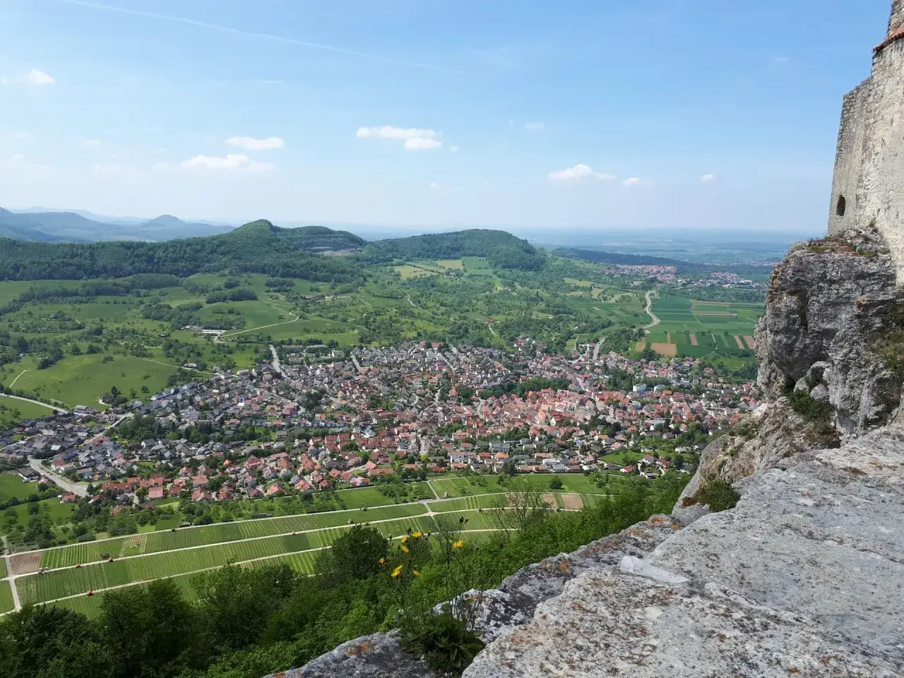 Blick von einer Felswand auf die Ruine Hofen und das darunterliegende Dorf mit Weinbergen und grünen Hügeln.
