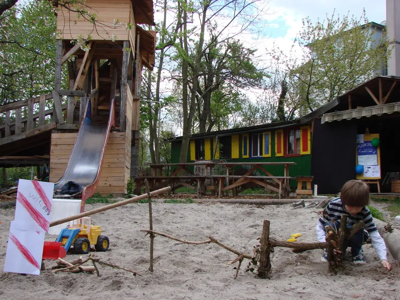 Ein Junge spielt auf dem abenteuerspielplatz stuttgart. Ein Holzturm mit Rutsche und ein bunter Bauwagen stehen im Sand.