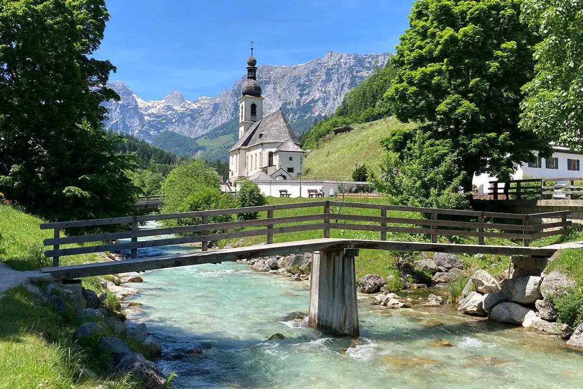 Idyllische Kirche mit Bergkulisse, ein Fluss fließt unter einer Holzbrücke hindurch. Perfekt für einen Kurztrip Deutschland.