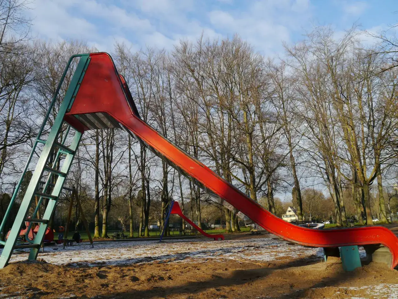 Ein rotes Flugzeug-Spielplatz-Gerüst mit Leiter und Rutsche steht im Sand. Im Hintergrund kahle Bäume und blauer Himmel.