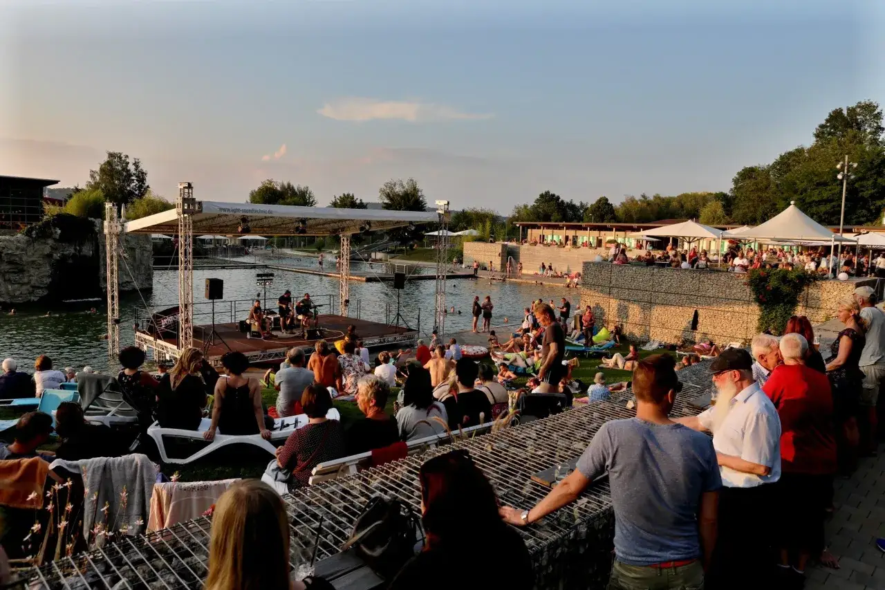 Sommerabend am Badesee Schorndorf: Menschen entspannen, baden und genießen ein Konzert am Wasser.