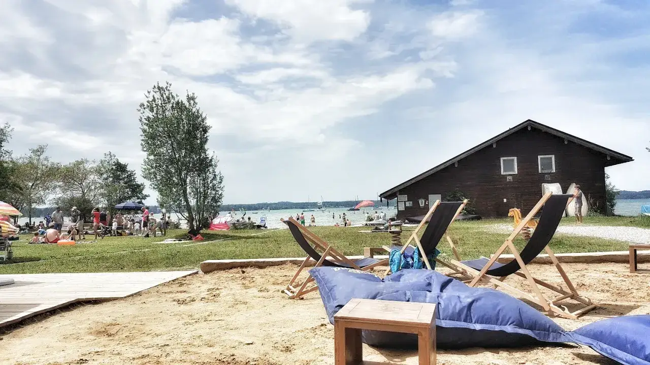 Sonniger Tag am Starnberger See mit vielen Familien und Kindern, die am Strand spielen und im Wasser planschen. Liegest&uuml;hle und Sitzs&auml;cke laden zum Entspannen ein.