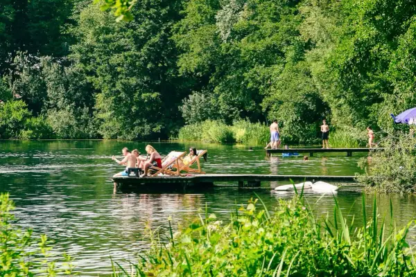 Menschen entspannen sich auf einem Floß und genießen die Sonne an einem Münchner See. Andere stehen auf einem Steg, einige baden im Wasser.