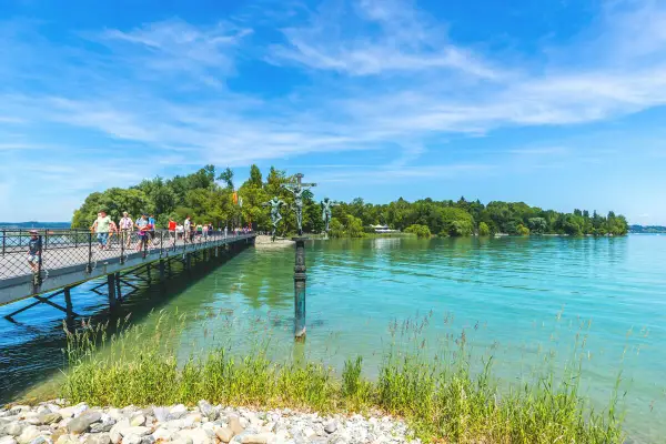 Menschen spazieren auf einer Brücke am Bodensee. Ein Denkmal mit Kreuzigungsgruppe steht im Wasser. Hier kann man in Konstanz viel unternehmen.